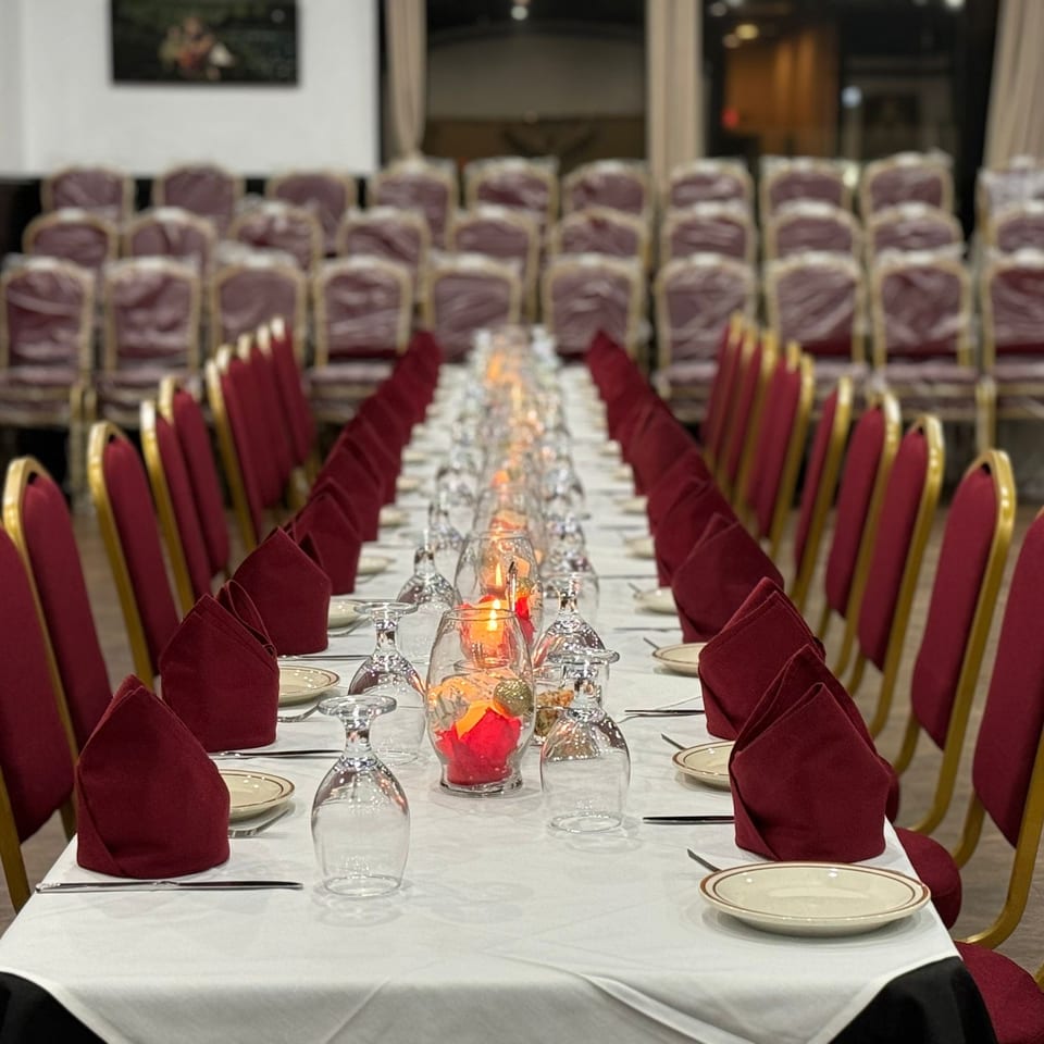 Long dining table set with white tablecloth, red napkins, plates, glasses, and candles, flanked by red chairs.