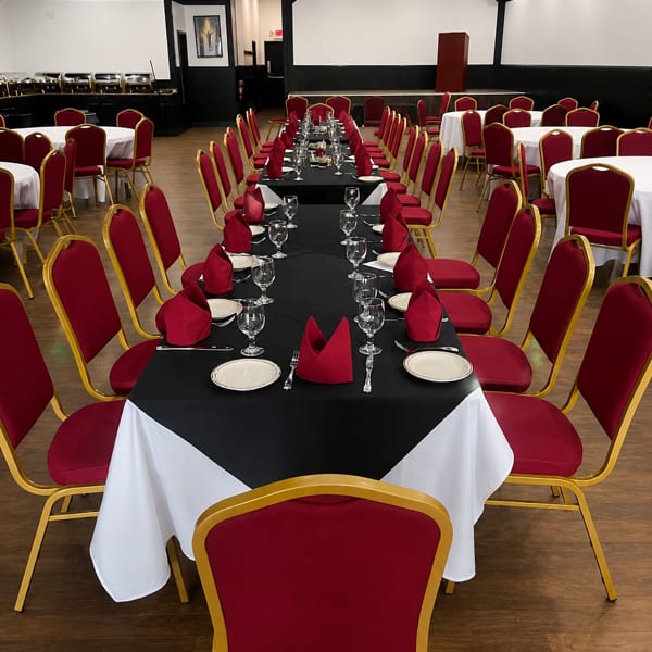Long dining tables with black and white tablecloths, red napkins, and gold-framed red chairs in a banquet hall.