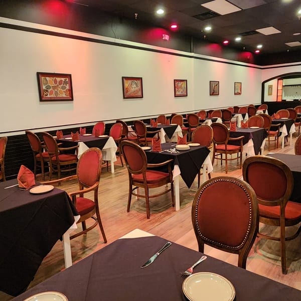 Empty restaurant dining room with black tablecloths, red chairs, and framed artwork on white walls.